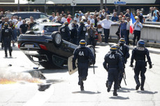 French riot police stand next to an overturned car as striking French taxi drivers demonstrate at Porte Maillot to block the traffic on the Paris ring road during a national protest against car-sharing service Uber, in Paris